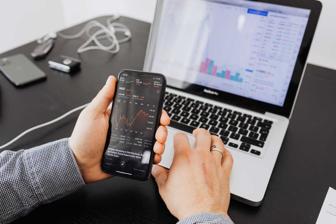 Person examines stock data on a smartphone and laptop indoors.